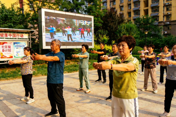 市民在社区健身广场跟随大屏幕进行健身操锻炼，政策宣传栏在一旁